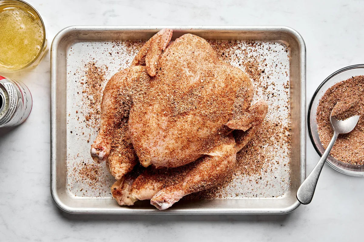 a whole raw chicken being generously seasoned on a baking sheet getting prepped for beer can chicken