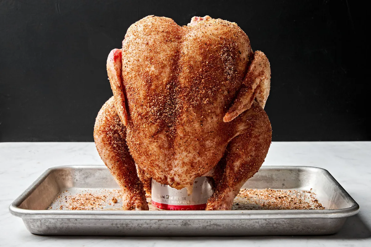 A whole raw chicken generously seasoned and being placed upright onto a beer can preparing to grill