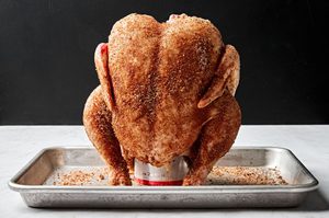A whole raw chicken generously seasoned and being placed upright onto a beer can preparing to grill