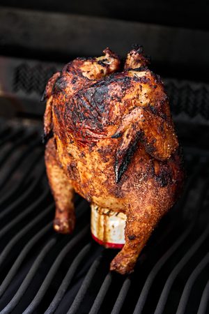 Perfectly seasoned whole beer can chicken cooking upright on a grill, skin turning crisp and golden.