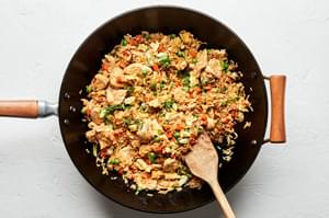 chicken fried rice being stirred in a wok with a wooden spoon with carrots, onions, peas, green onions, eggs & soy sauce