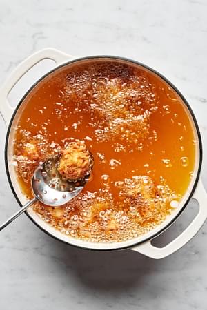 homemade chicken nuggets being fried in a large pot of vegetable oil and removed with a metal slotted spoon