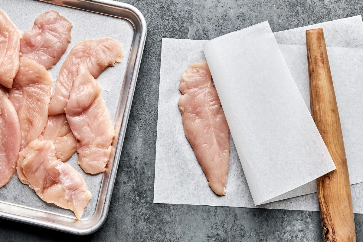 raw chicken tenders being pounded flat between parchment paper to make chicken satay