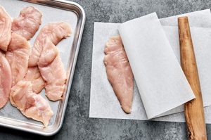 raw chicken tenders being pounded flat between parchment paper to make chicken satay