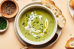 a bowl of homemade cream of broccoli soup sprinkled with chives and garnished with a swirl of cream served with crusty bread