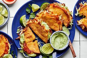 4 crispy chicken tacos on a plate topped with hot sauce and shredded lettuce, served with lime wedges and avocado sauce