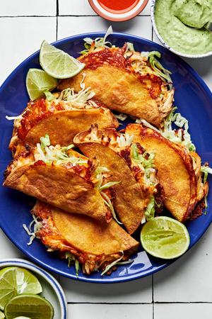 6 crispy chicken tacos on a plate topped with hot sauce and shredded lettuce, served with lime wedges and avocado sauce