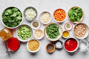ingredients for minestrone soup in prep bowls: stock, pasta, vegetables, spices, cannellini beans, fresh basil and parmesan