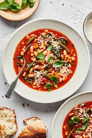 a bowl of homemade minestrone soup topped with parmesan and fresh basil surrounded by parmesan, basil and bread for serving