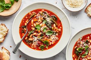 a bowl of homemade minestrone soup topped with parmesan and fresh basil surrounded by parmesan, basil and bread for serving
