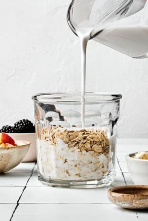 coconut milk being poured over oats in a glass jar to make overnight oats