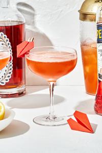 a paper plane cocktail in a coupe glass with a small paper airplane attached to the side of the glass