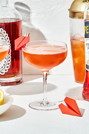 a paper plane cocktail in a coupe glass with a small paper airplane attached to the side of the glass