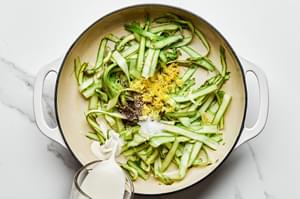 heavy cream being added into a skillet with olive oil, asparagus ribbons, garlic, pepper, lemon zest & salt