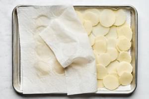 Thinly sliced potatoes being pat completely dry between paper towels.