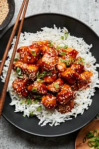 a bowl of homemade sesame chicken on top of white rice surrounded by bowl of green onions, sesame seeds for garnishing