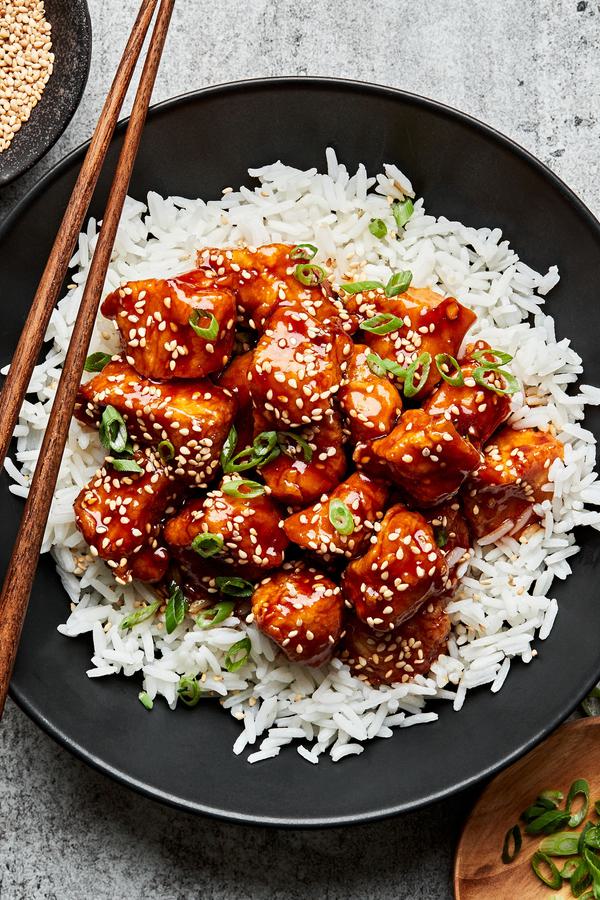 a bowl of homemade sesame chicken on top of white rice surrounded by bowl of green onions, sesame seeds for garnishing