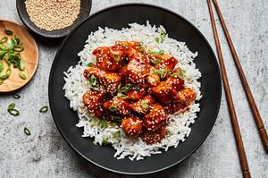 a bowl of homemade sesame chicken on top of white rice surrounded by bowl of green onions, sesame seeds for garnishing