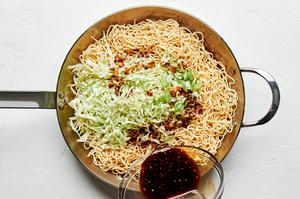 cooked chow mein noodles , soy sauce mixture, green onion, and cabbage being cooked in sesame oil in a skillet