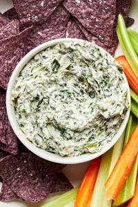a bowl of homemade spinach dip surrounded by blue corn tortilla chips, carrot sticks and celery sticks for dipping