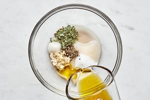 olive oil being poured into a small bowl with garlic, vinegar, sugar, basil, salt, and pepper