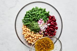 homemade dressing being poured over top of green beans, kidney beans, garbanzo beans, red onion and parsley