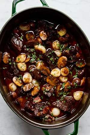 a pot of beef bourguignon made with mushrooms, pear onions, carrots, onion, tomato paste, red wine, and spices