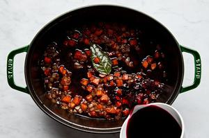 red wine being poured into a dutch oven with onion, carrot, garlic, tomato paste, bay leaf, thyme and flour
