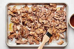 cooked carnitas on a baking sheet being brushed with reserved liquid from the dutch oven pot