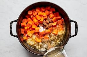 carrots, spices, and vegetable stock being added to a soup pot with cooked onion and garlic