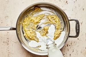 milk being added to a skillet with butter, shallots, garlic and flour to make a roux