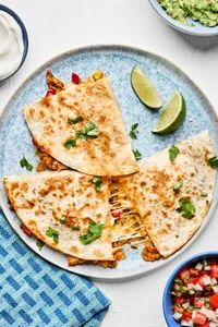 a sliced chicken quesadilla on a plate surrounded by sour cream, guacamole and pico de gallo for serving