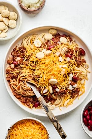 a bowl of Cincinnati chili served on spaghetti noodles topped with cheese, kidney beans and crackers surrounded by toppings