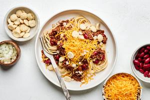 a bowl of Cincinnati chili served on spaghetti noodles topped with cheese, beans and oyster crackers surrounded by toppings
