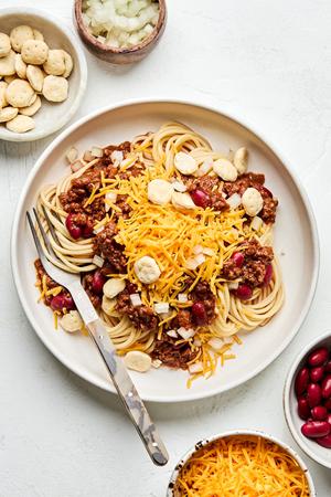 a bowl of Cincinnati chili served on spaghetti noodles topped with cheese, kidney beans and crackers surrounded by toppings