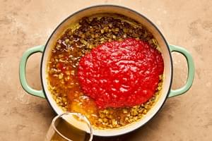 crushed tomatoes and chicken stock being added to a pot with cooked onion and spices