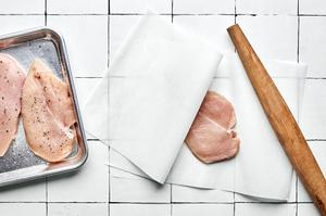 chicken breast being pounded thin with a rolling pin between 2 pieces of parchment paper