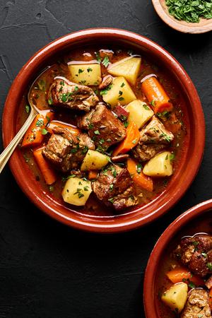 a bowl of pork stew made with potatoes, carrots, onion, carrots, bacon, spices and tomato paste sprinkled with parsley