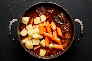 potatoes and carrots being added to a pot of pork stew with bacon
