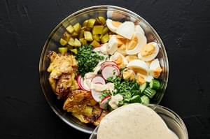 dressing being poured over smashed potatoes, boiled eggs, pickles, cucumber, radish, parsley and chives