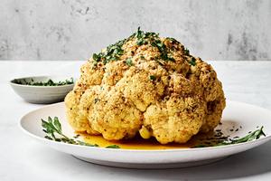 a whole roasted cauliflower on a serving plate seasoned with garlic, italian, salt, paprika & pepper & topped with Parmesan
