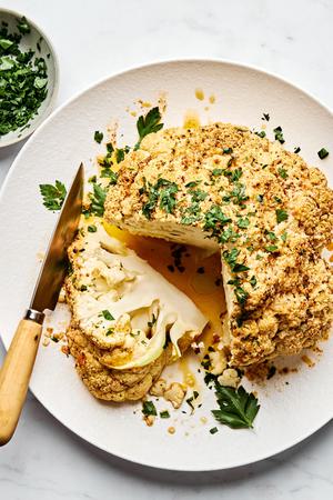 a whole roasted cauliflower topped with Parmesan and parsley being sliced into wedges on a serving plate