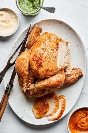 a whole roasted chicken on a serving plate with a carving knife served with romesco sauce, pesto and garlic aioli for dipping
