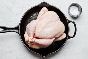 a whole raw chicken in a cast iron skillet being seasoned with salt