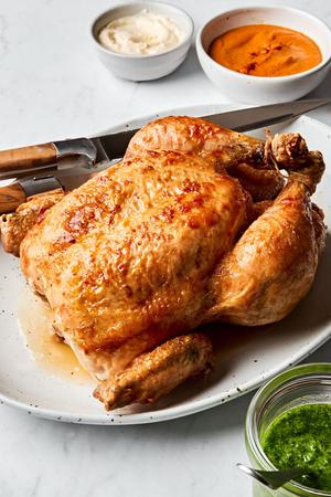a whole roasted chicken on a serving plate with a carving knife served with romesco sauce, pesto and garlic aioli for dipping