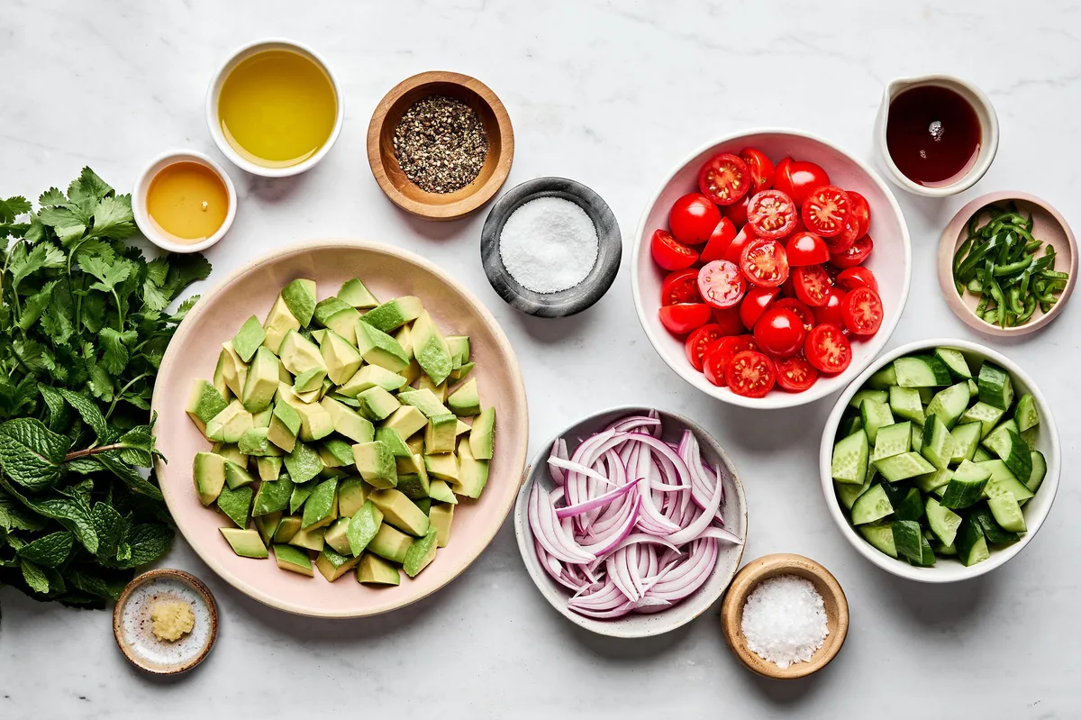 Ingredients for avocado salad including avocados, cherry tomatoes, cucumbers, red onion, herbs, vinegar, oil, and spices.