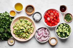 Ingredients for avocado salad including avocados, cherry tomatoes, cucumbers, red onion, herbs, vinegar, oil, and spices.