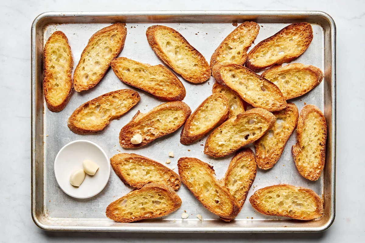 toasted, oiled baguette slices on a baking sheet being rubbed with garlic cloves to make easy homemade bruschetta