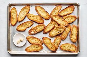 toasted, oiled baguette slices on a baking sheet being rubbed with garlic cloves to make easy homemade bruschetta
