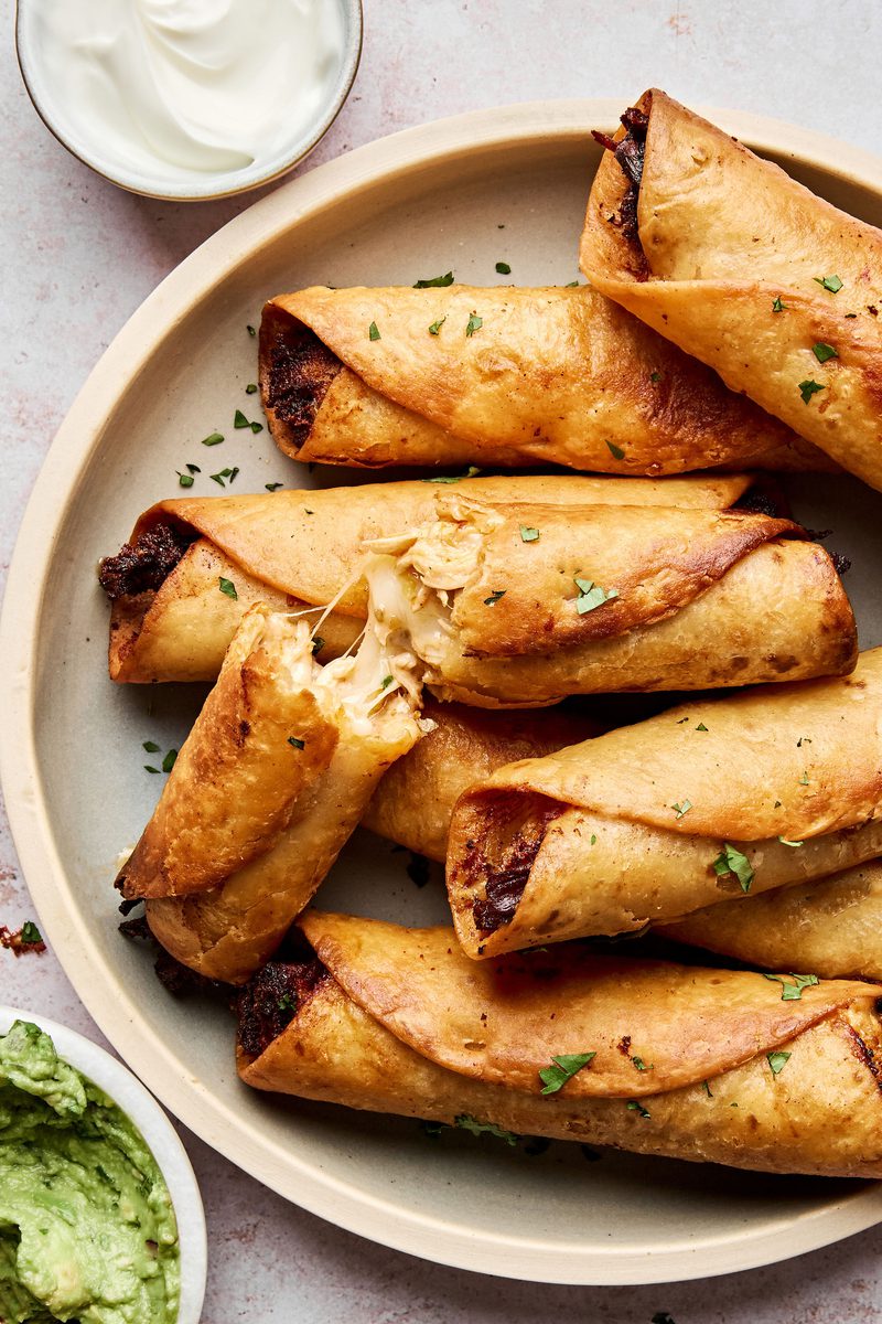 A plate of golden, crispy homemade chicken flautas served with sour cream and guacamole on the side.